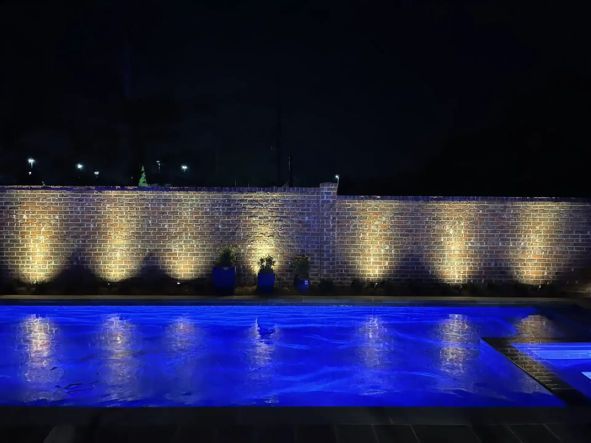 Low-voltage landscape lights washing a brick wall beside a pool at night